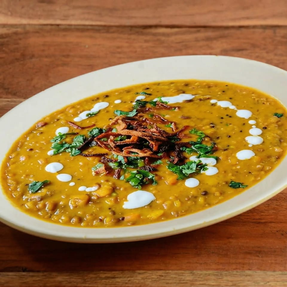 A bowl of yellow lentil dal with fried onions, cilantro, and cream on a wooden table.