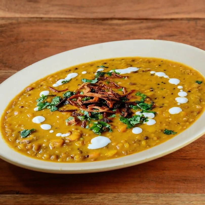A bowl of yellow lentil dal with fried onions, cilantro, and cream on a wooden table.
