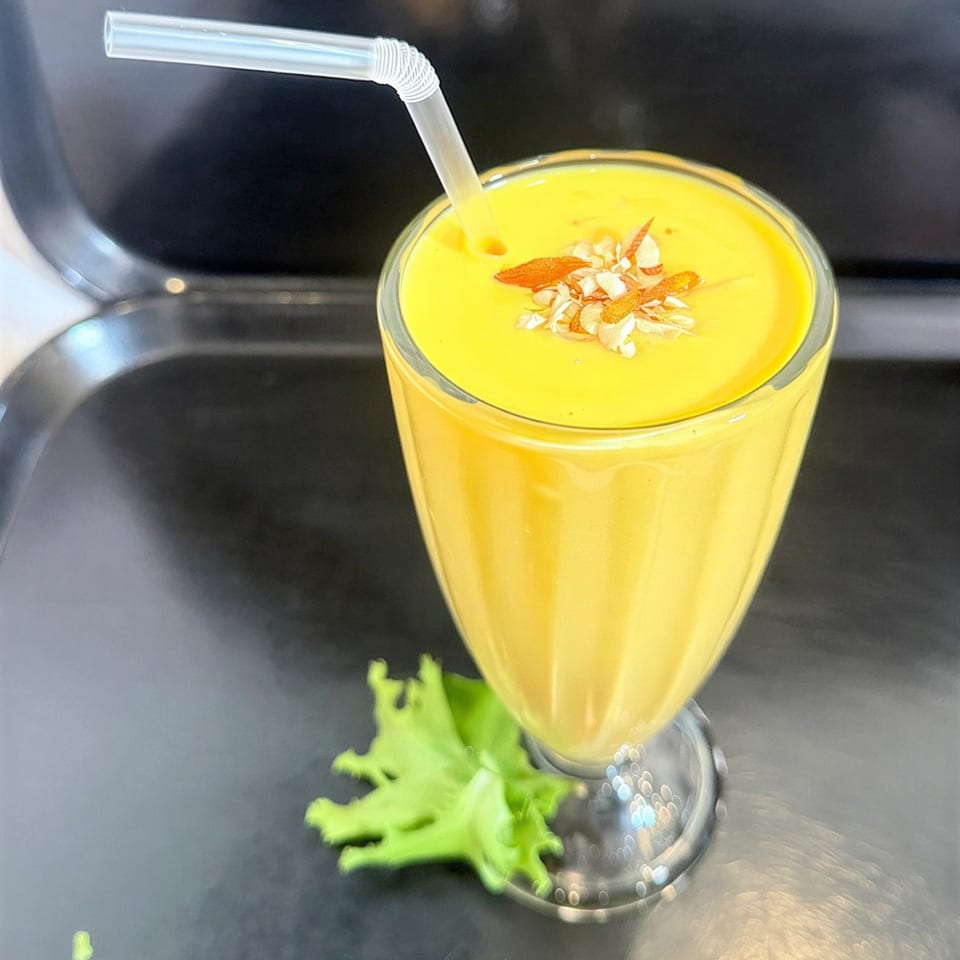 A vibrant yellow mango lassi in a glass, garnished with almonds and saffron, with a straw.