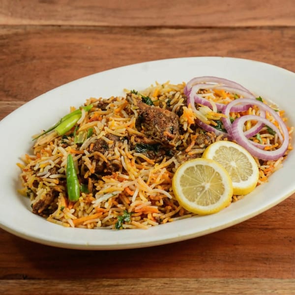 A white oval plate filled with chicken biryani, garnished with red onion rings, green chilies, and lemon slices.