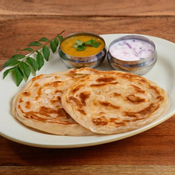 Two flaky parathas with yellow dal and raita in steel bowls, garnished with curry leaves.