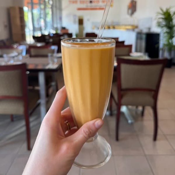 A hand holds a tall glass of orange-colored lassi with a straw in a restaurant setting.