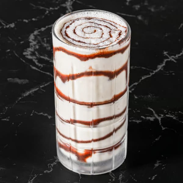 Tall glass of white milkshake with red and brown syrup swirls, on a dark marble surface.