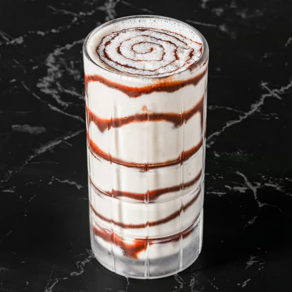 Tall glass of white milkshake with red and brown syrup swirls, on a dark marble surface.