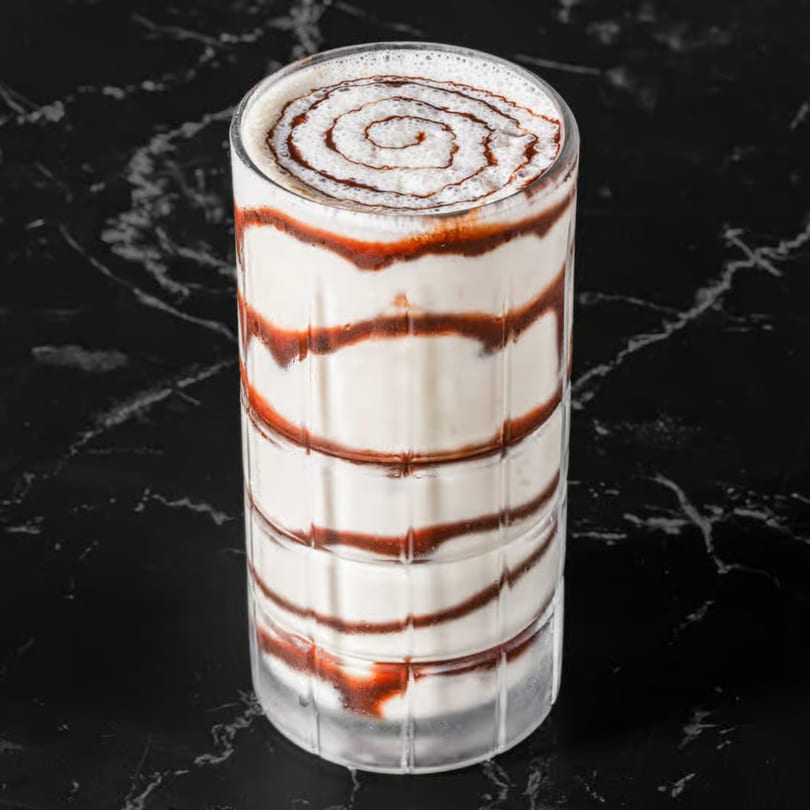 Tall glass of white milkshake with red and brown syrup swirls, on a dark marble surface.