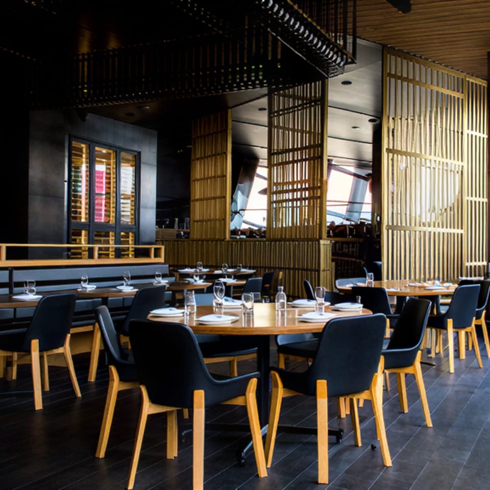 Modern restaurant interior with dark wood floors, round tables, black chairs, and golden slatted screens.
