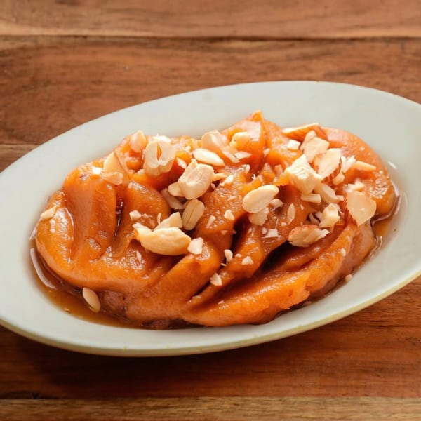 A white oval plate holds a serving of orange-brown dessert topped with chopped nuts, on a wooden table.