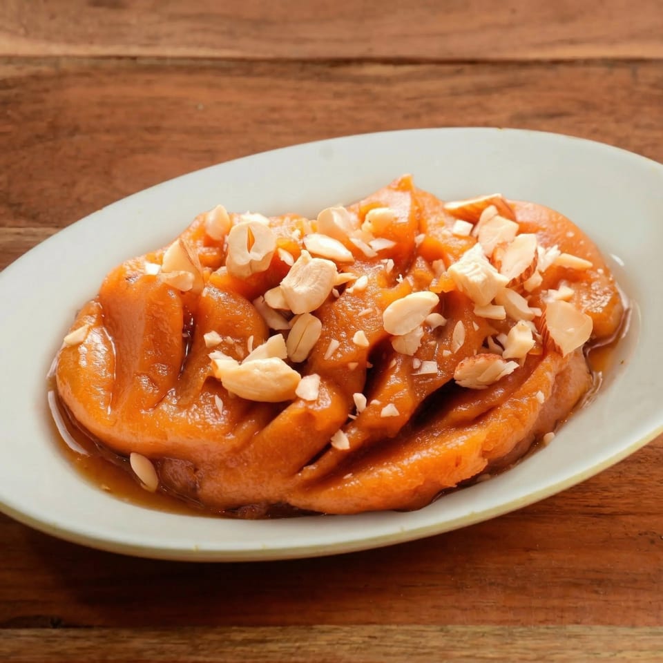A white oval plate holds a serving of orange-brown dessert topped with chopped nuts, on a wooden table.