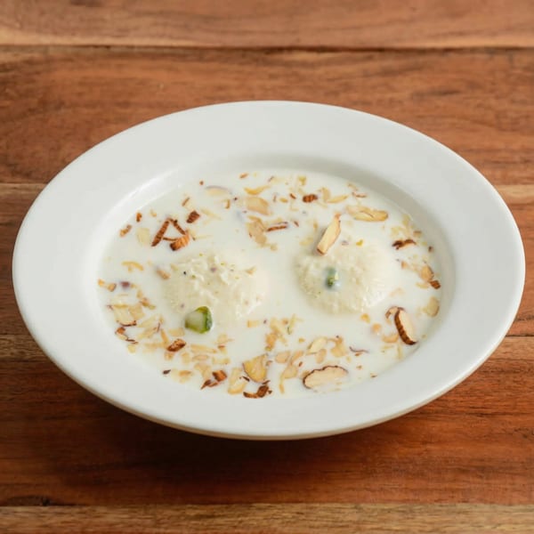 Two rasgullas in creamy milk, garnished with sliced almonds and pistachios, in a white bowl.