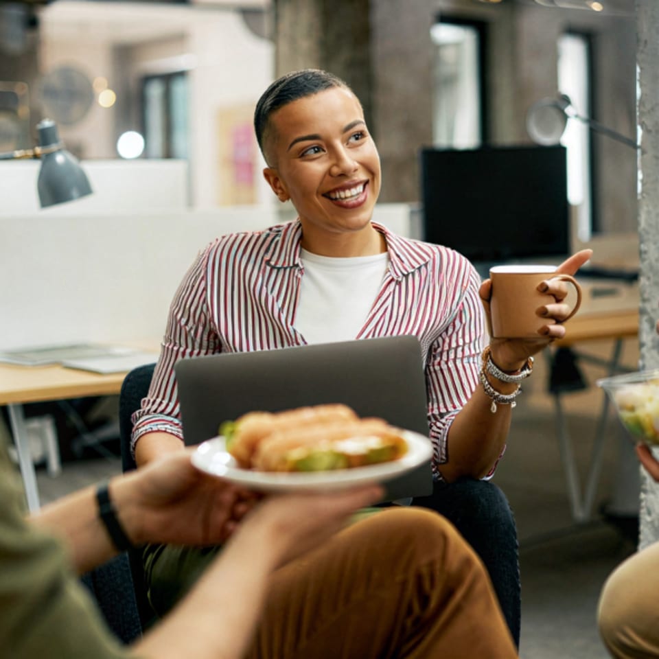 Smiling woman with short hair holding a coffee mug, looking at a plate of sandwiches.